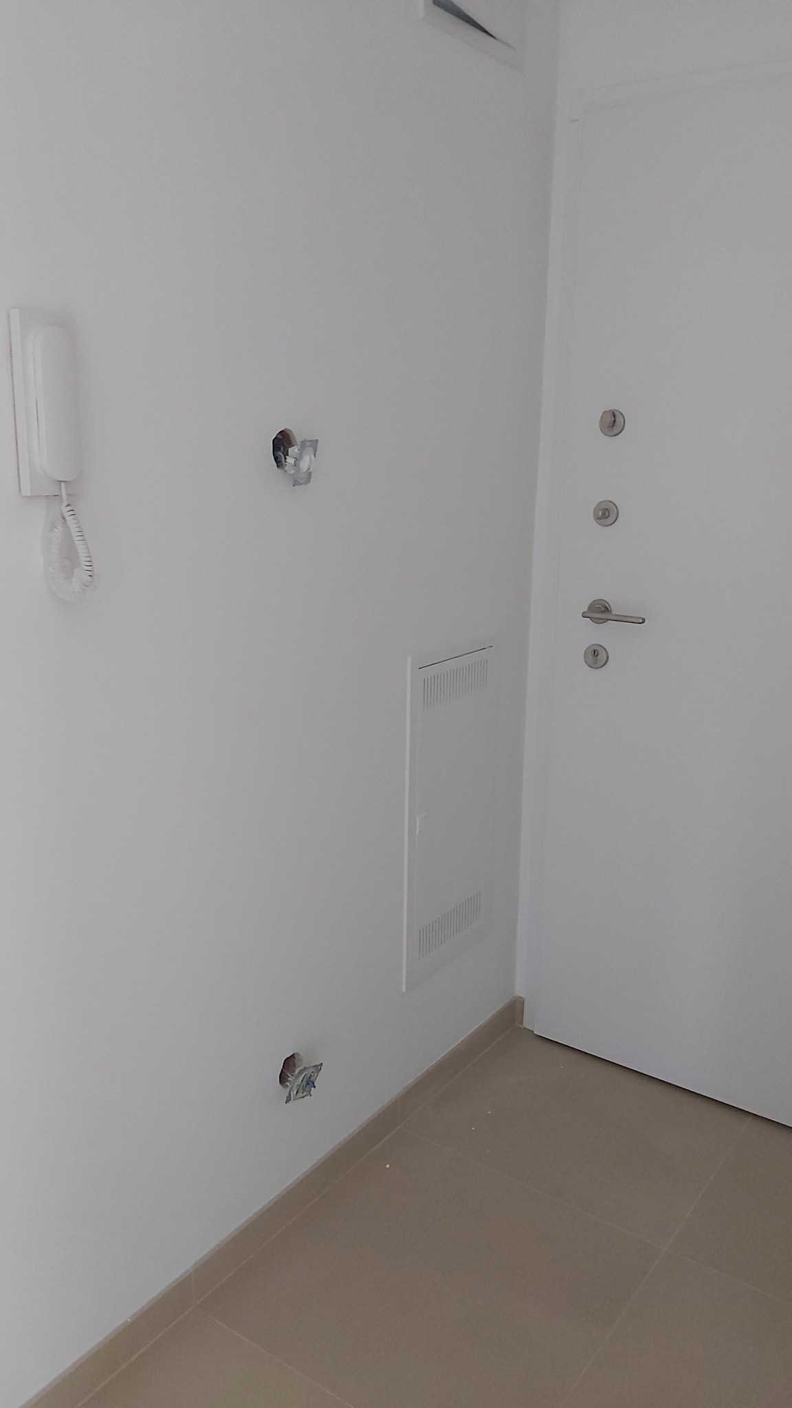 Wnętrze mieszkania w trakcie prac wykończeniowych: białe ściany z widocznymi puszkami elektrycznymi bez osprzętu, domofon na ścianie, obok białe drzwi wejściowe, na podłodze beżowe płytki.