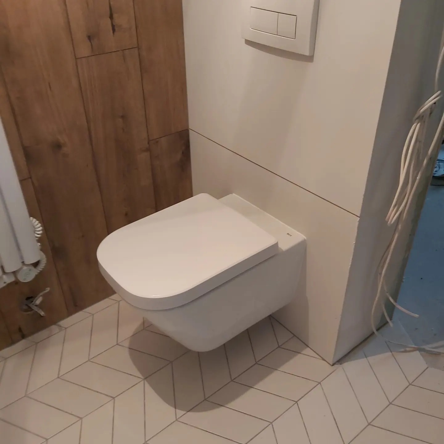 Nowoczesna, podwieszana toaleta z białą deską, zamontowana na białej ścianie z drewnianymi panelami po lewej stronie i geometryczną podłogą w łazience po remoncie.