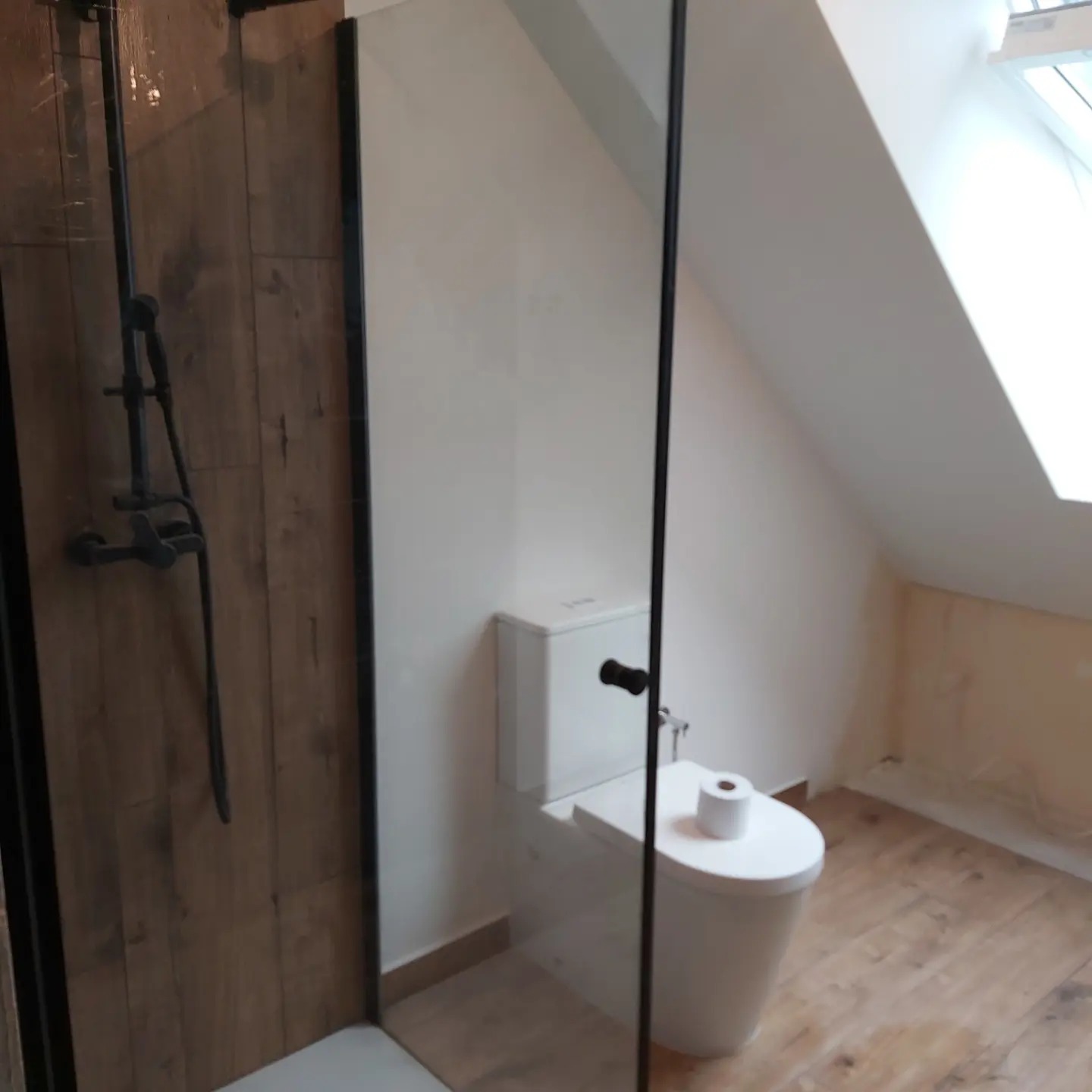 Nowoczesna łazienka na poddaszu z kabiną prysznicową typu walk-in, drewnopodobnymi płytkami i białą toaletą z rolką papieru.