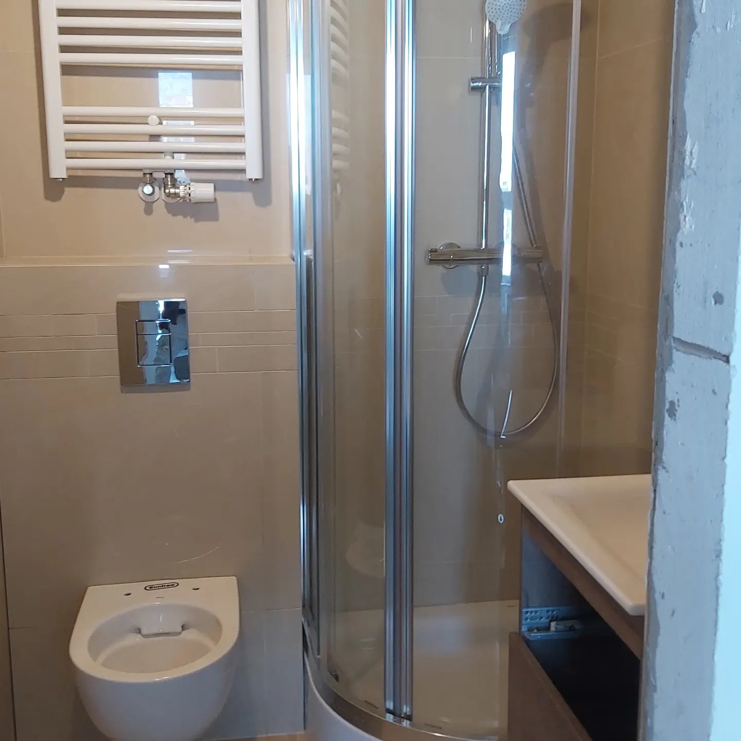 Nowoczesna łazienka z beżowymi płytkami, kabiną prysznicową z przezroczystego szkła, podwieszaną toaletą, grzejnikiem drabinkowym i szafką z umywalką, widoczny fragment niezakończonych prac...