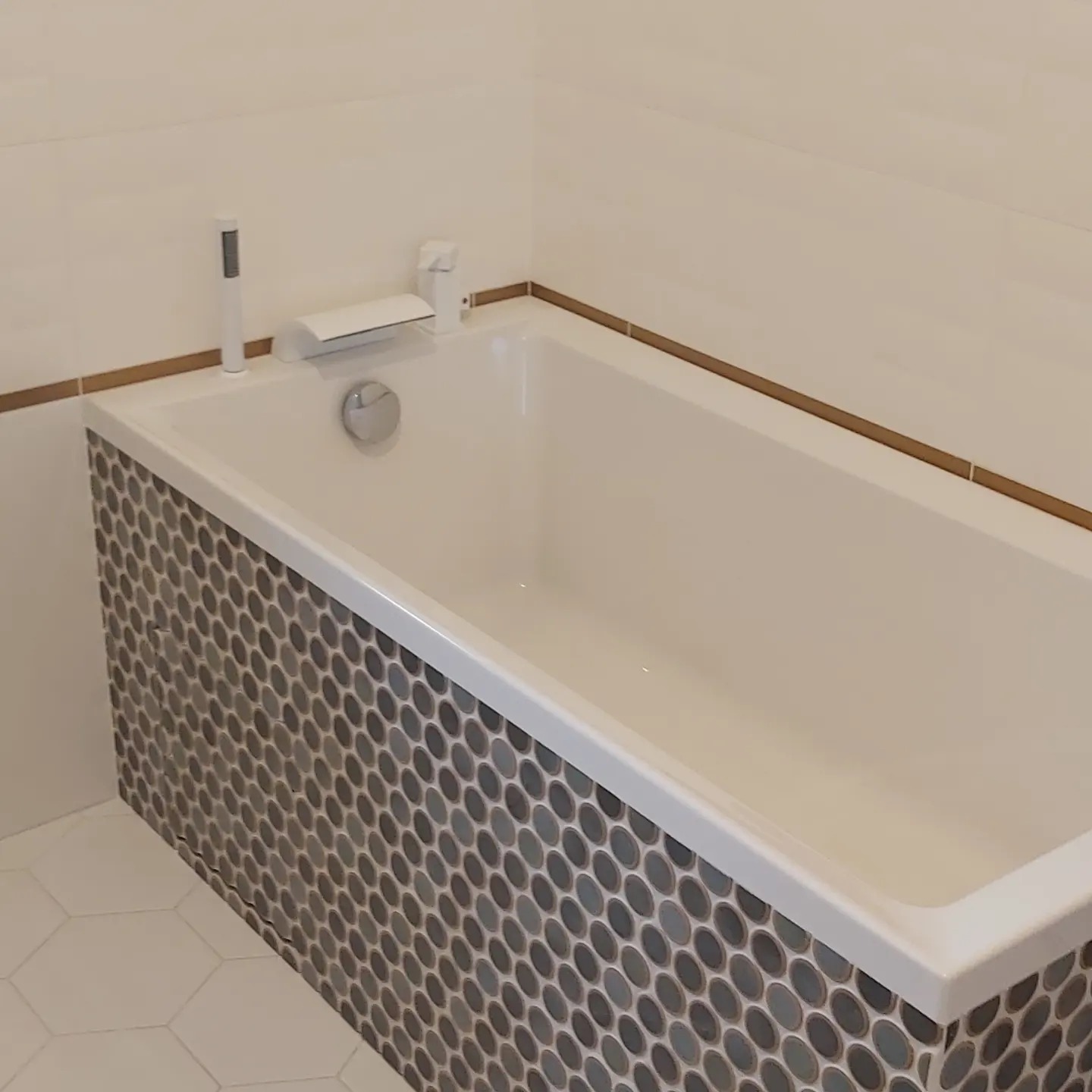 Nowoczesna, biała wanna obudowana mozaiką w odcieniach szarości, z minimalistyczną armaturą i białymi, prostokątnymi płytkami na ścianie, wykończonymi brązową listwą. Na podłodze widoczne białe...