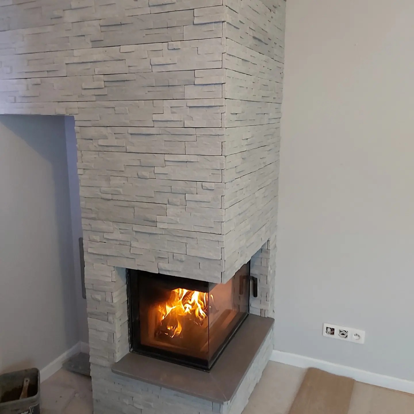 Obudowa kominka wyłożona szarym kamieniem dekoracyjnym o fakturze łupka, z widocznym paleniskiem i płomieniami, oraz fragmentem ściany z gniazdkami elektrycznymi.