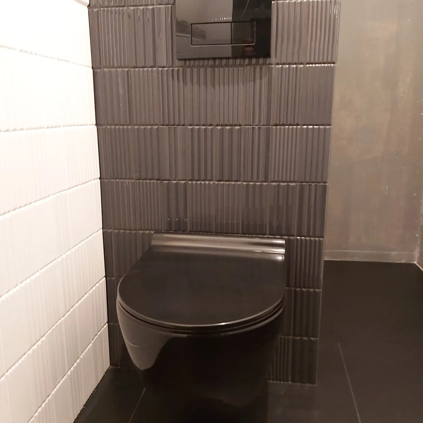 Nowoczesna łazienka z wiszącą toaletą w kolorze czarnym, otoczoną ścianami wyłożonymi pionowymi, teksturowanymi płytkami w odcieniach szarości i bieli.