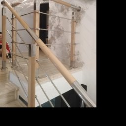 Dod-met - Nowoczesna balustrada schodowa z drewnianymi słupkami i metalowymi poprzeczkami, częściowo zabezpieczona folią ochronną, zamontowana na marmurowych schodach wewnątrz budynku.