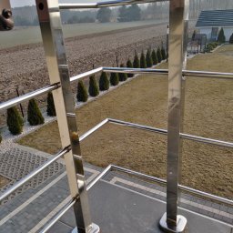 Dod-met - Błyszcząca, metalowa konstrukcja zabezpieczająca taras z widokiem na pole i rząd tuj, uchwycona w słoneczny dzień.