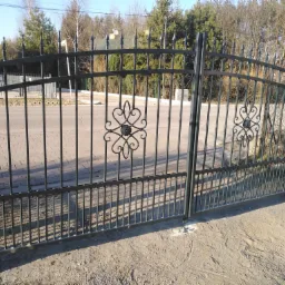 Czarna, metalowa brama wjazdowa z ozdobnymi elementami w kształcie serc i kwiatów, widoczna z bliska na tle ulicy i drzew.