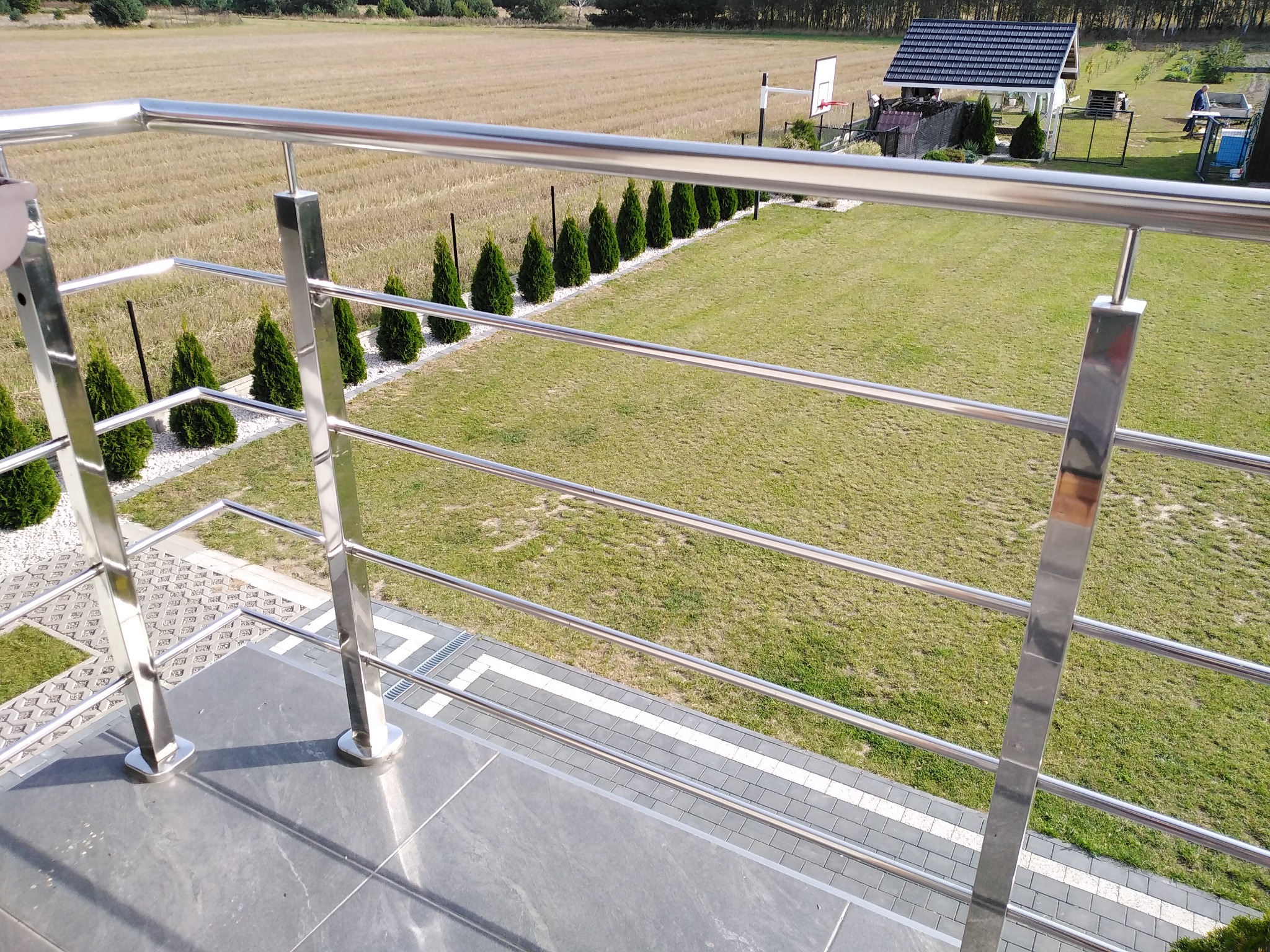 Błyszcząca, metalowa balustrada z widokiem na zadbany ogród z tujami i pole w tle, uchwycona z perspektywy balkonu wykończonego szarą płytką.