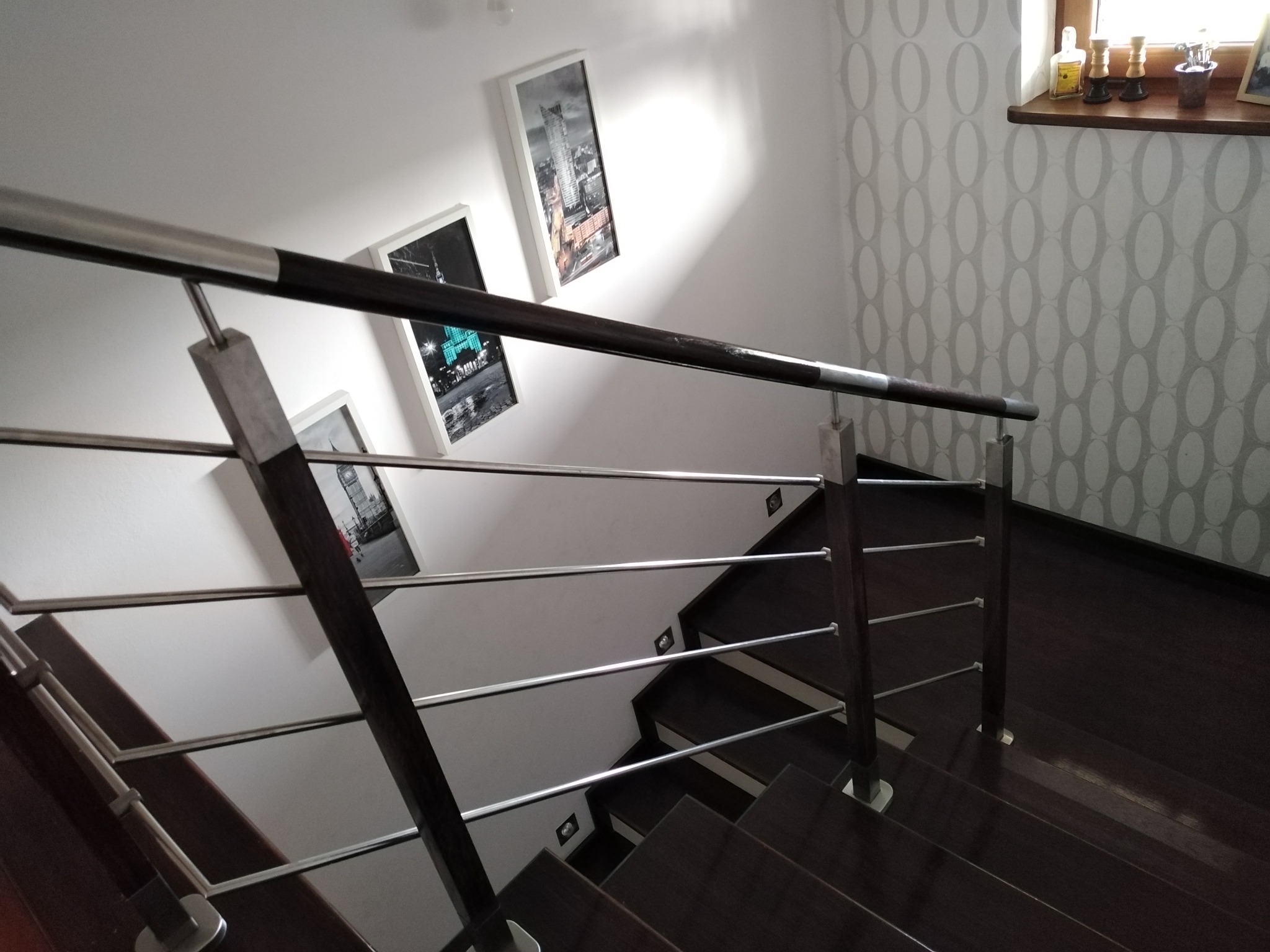Nowoczesne schody z ciemnego drewna z balustradą z elementami stali nierdzewnej, trzy obrazy w białych ramkach na ścianie, wzorzysta tapeta w tle.