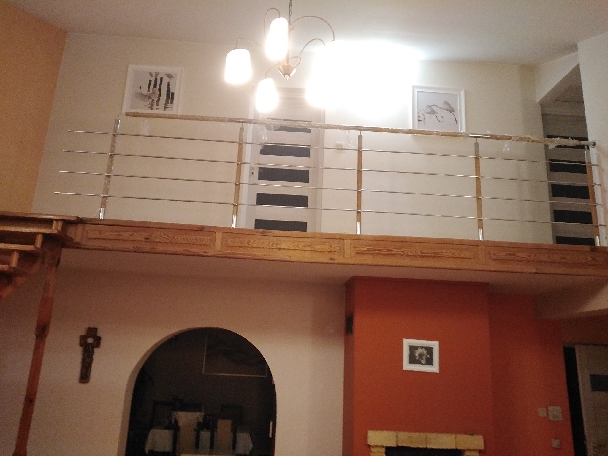 Wnętrze domu z antresolą zabezpieczoną nowoczesną balustradą ze stali nierdzewnej z poziomymi prętami, widok od dołu na drewnianą konstrukcję antresoli i dekoracje ścienne.