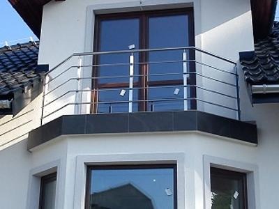 Zewnętrzna balustrada balkonowa ze stali nierdzewnej z poziomymi rurkami, zamontowana na balkonie z ciemnymi płytkami, widok z dołu na tle białej elewacji budynku z ciemną dachówką.