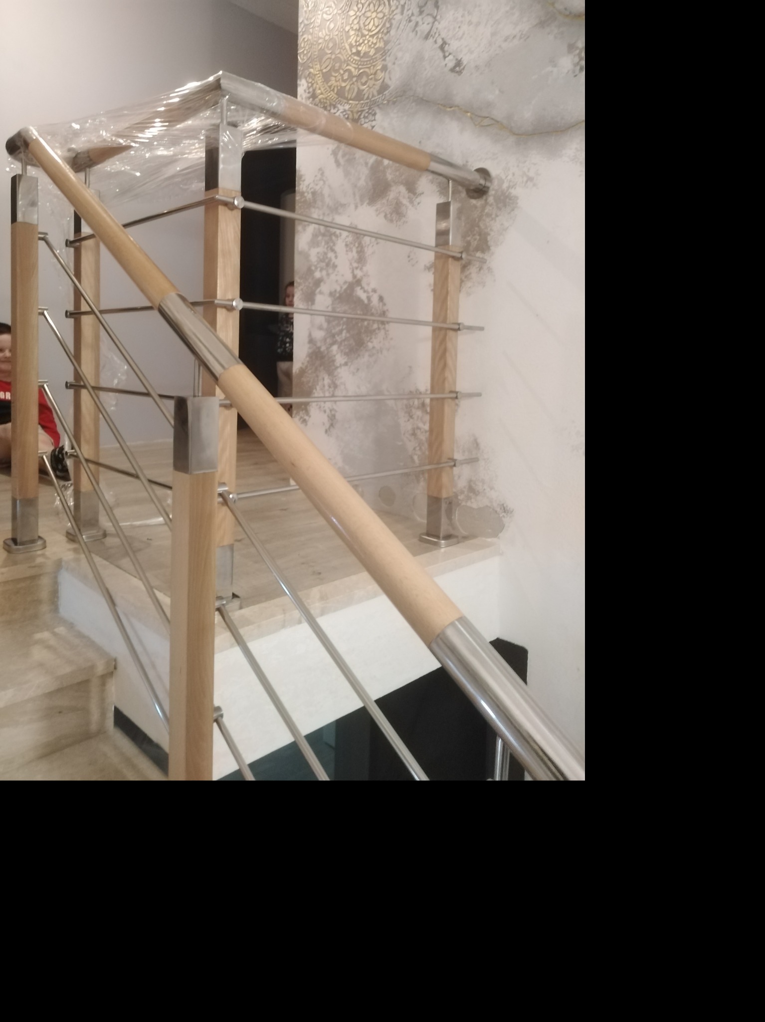 Nowoczesna balustrada schodowa z drewnianymi słupkami i metalowymi poprzeczkami, częściowo zabezpieczona folią ochronną, zamontowana na marmurowych schodach wewnątrz budynku.