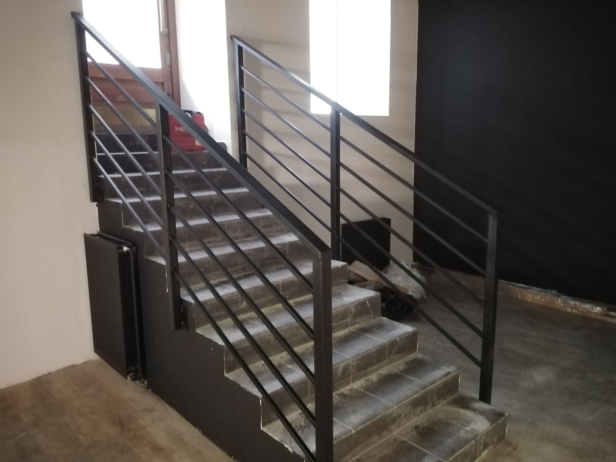 Schody z czarną, minimalistyczną barierką z poziomych prętów, widok z dołu, w tle biała ściana, okno i czarna ściana.