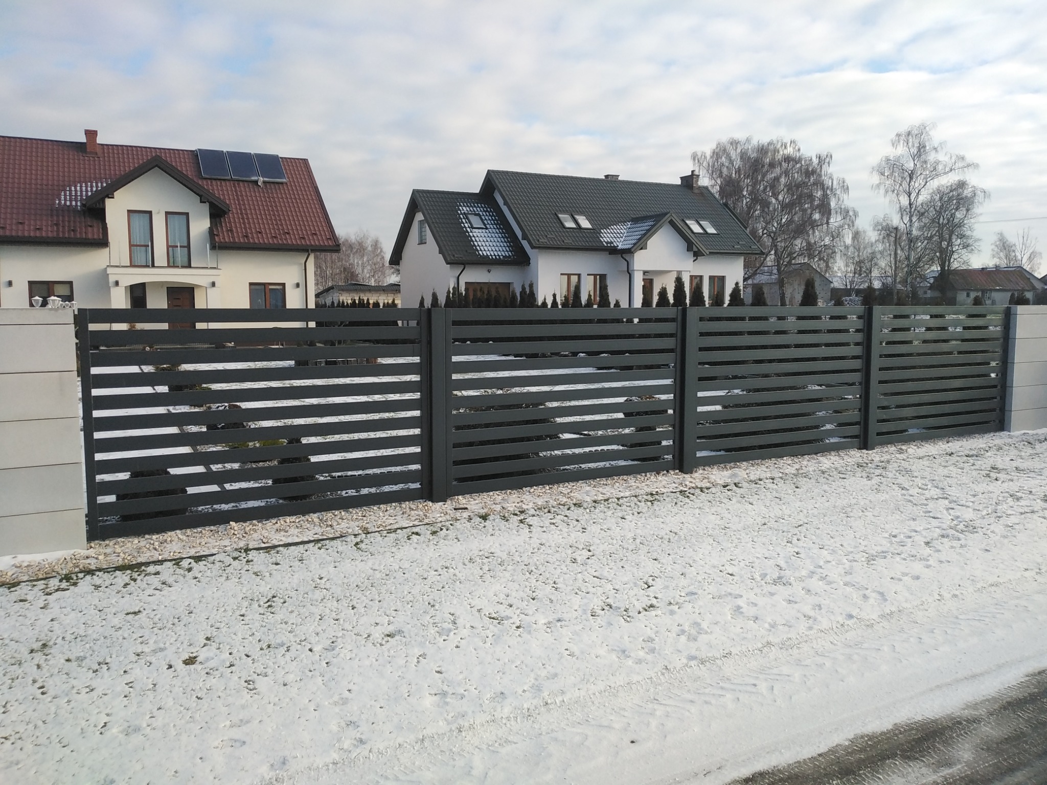 Nowoczesne, poziome przęsła w kolorze grafitowym, oddzielające posesję od drogi pokrytej śniegiem; w tle dwa białe domy z ciemnymi dachami.