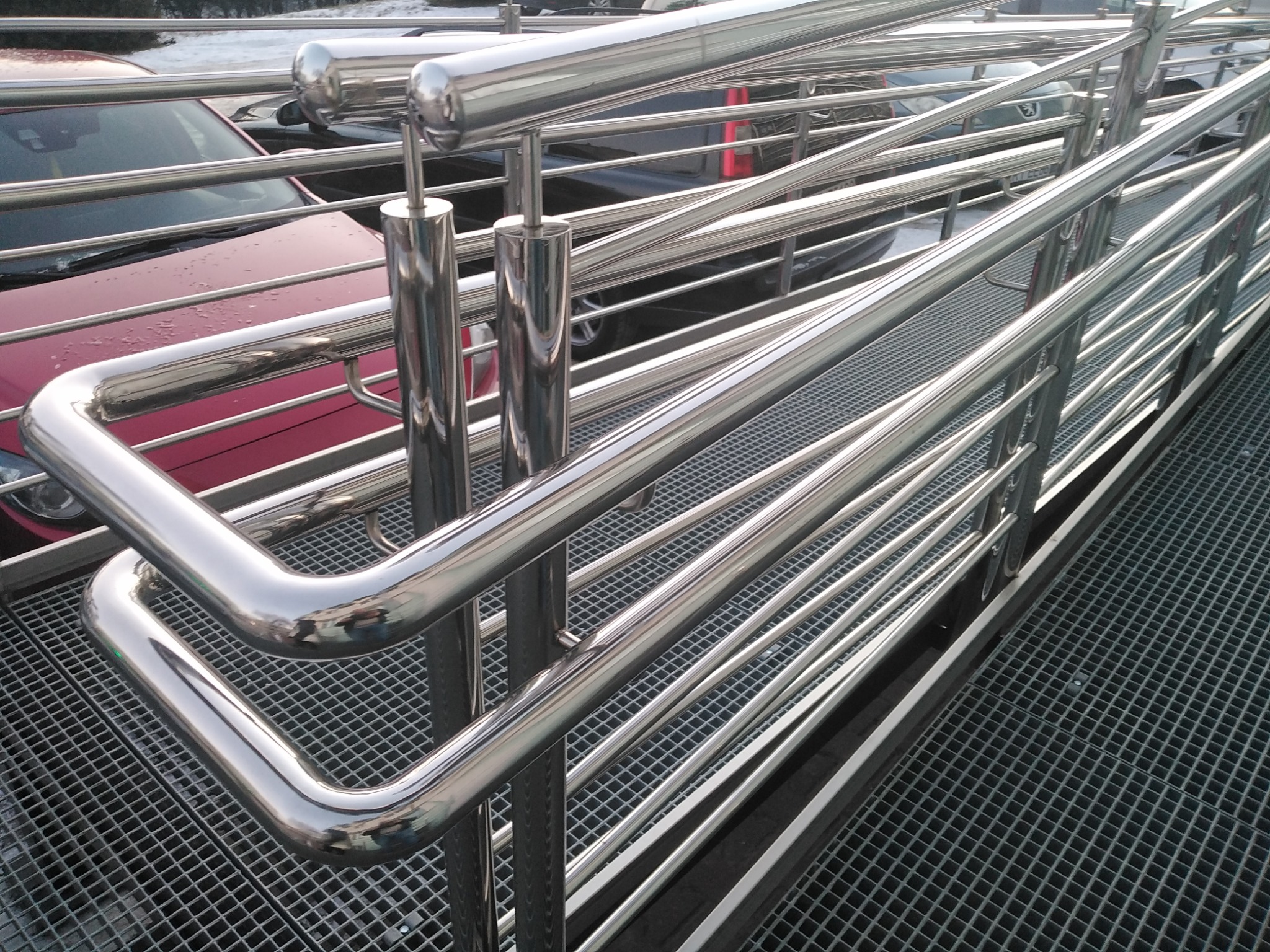 Rampa dla wózków inwalidzkich z metalową konstrukcją i poręczami ze stali nierdzewnej, widok z boku na tle zaparkowanych samochodów.