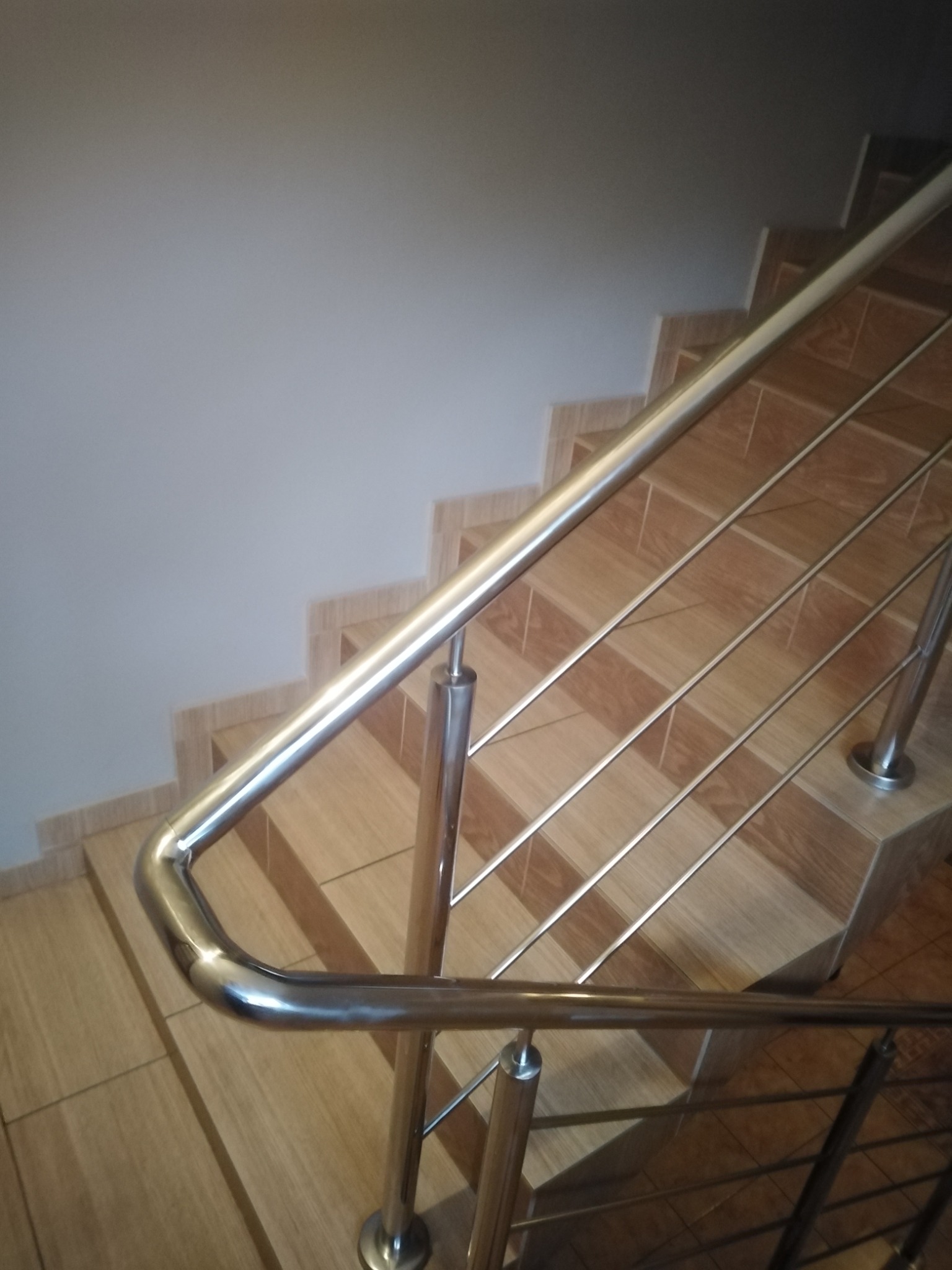 Metalowa poręcz schodowa z wypełnieniem prętami, zamontowana na schodach wyłożonych płytkami imitującymi drewno, widok z góry.