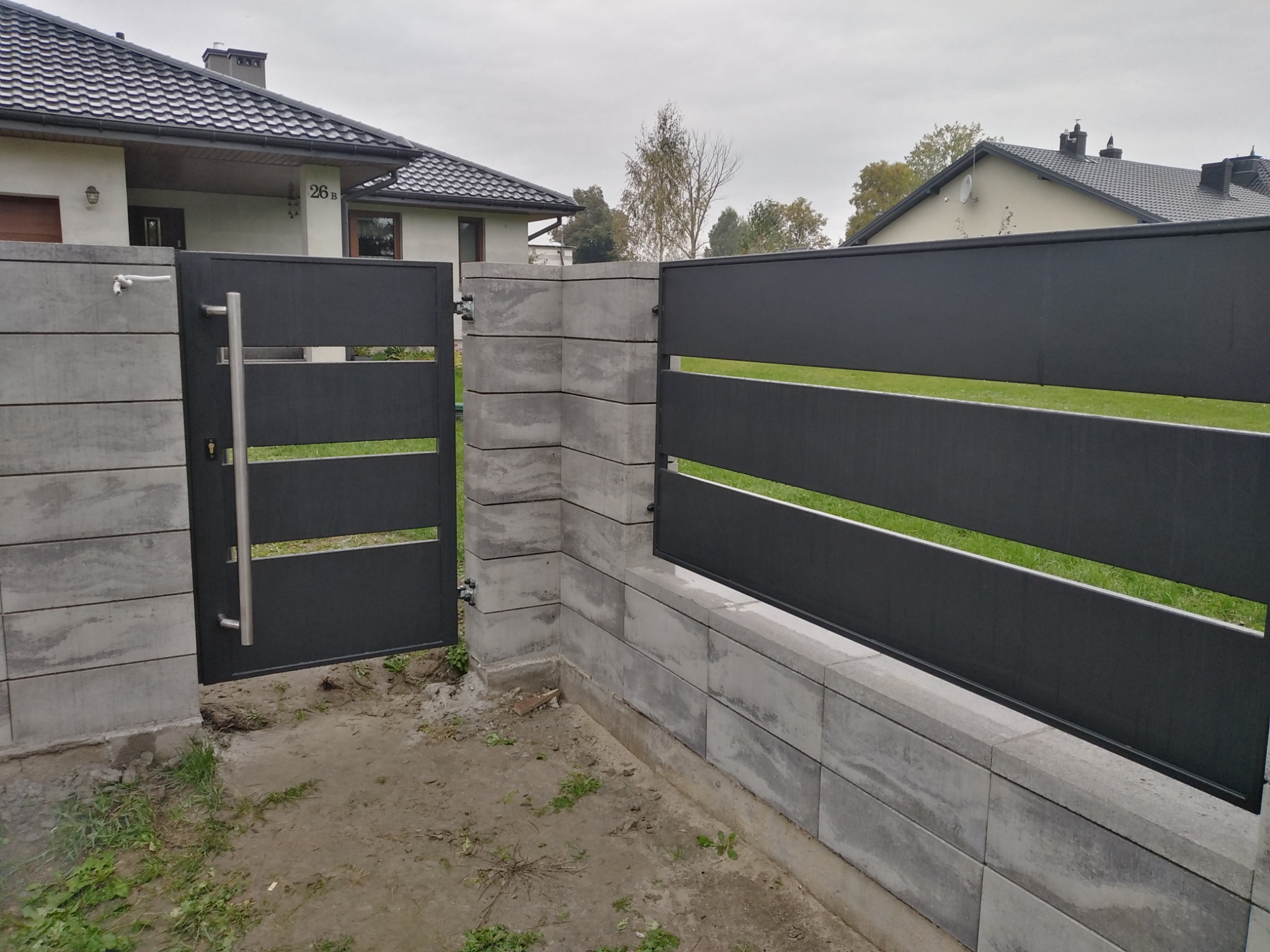 Nowoczesna, pozioma, metalowa brama wjazdowa w kolorze antracytowym, z uchwytem ze stali nierdzewnej, zamontowana na murku z szarej kostki betonowej, z widokiem na dom z ciemną dachówką