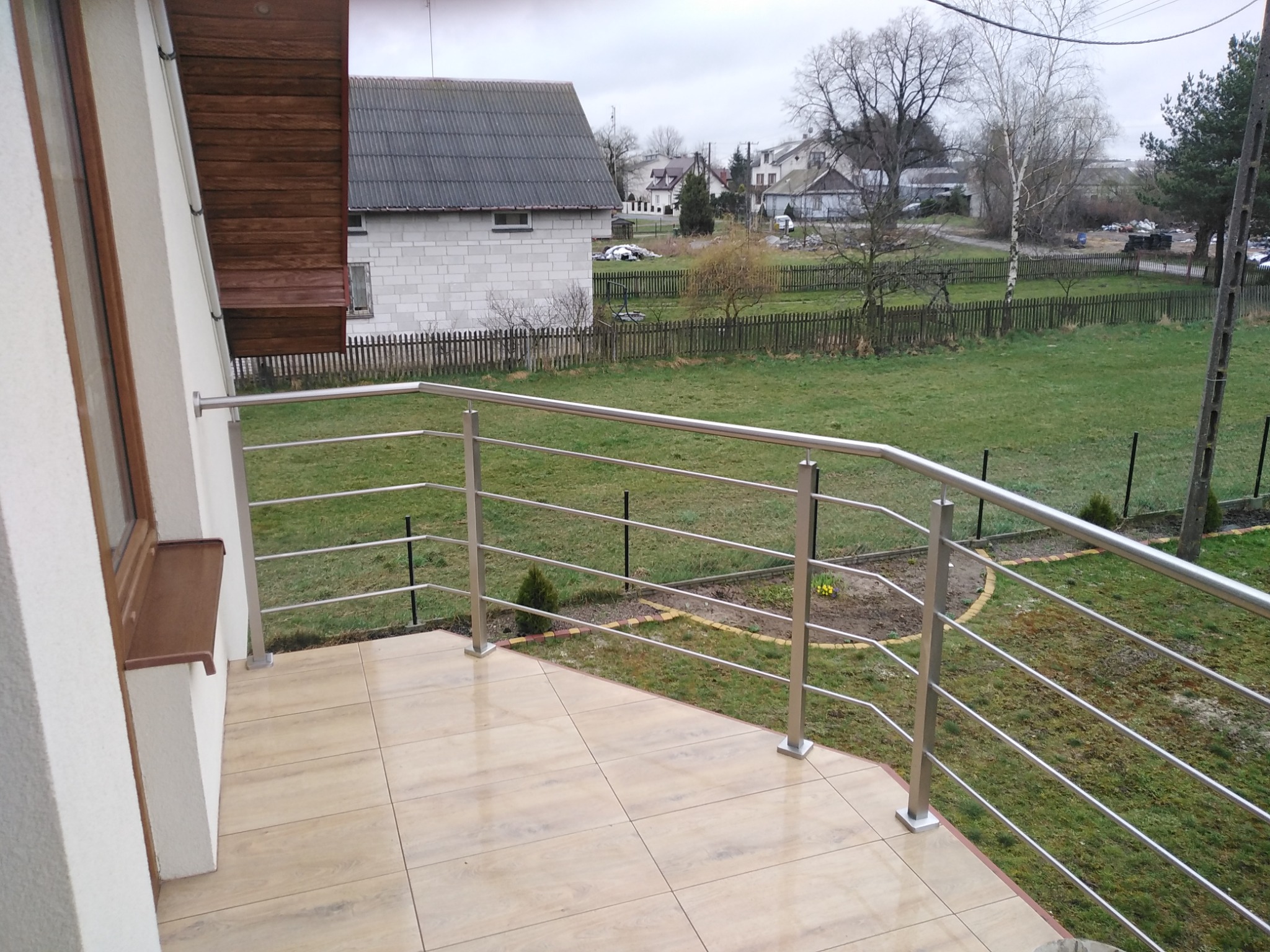 Balkon z jasnymi płytkami i nowoczesną, metalową barierką z widokiem na ogród i zabudowania w tle.