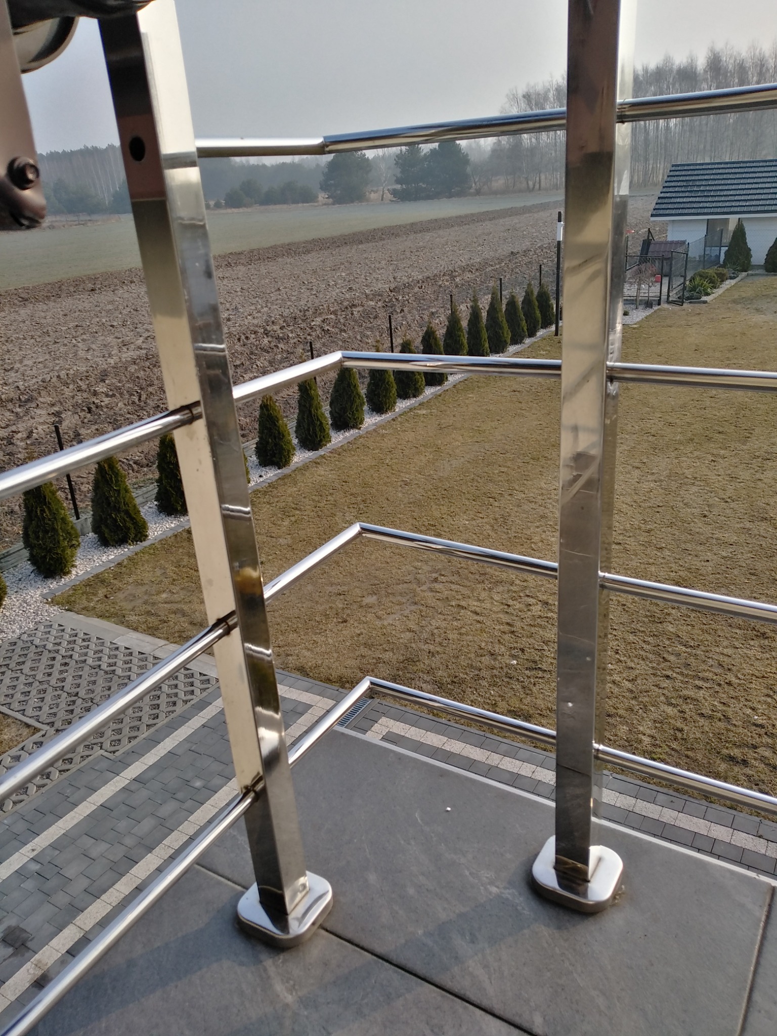 Błyszcząca, metalowa konstrukcja zabezpieczająca taras z widokiem na pole i rząd tuj, uchwycona w słoneczny dzień.