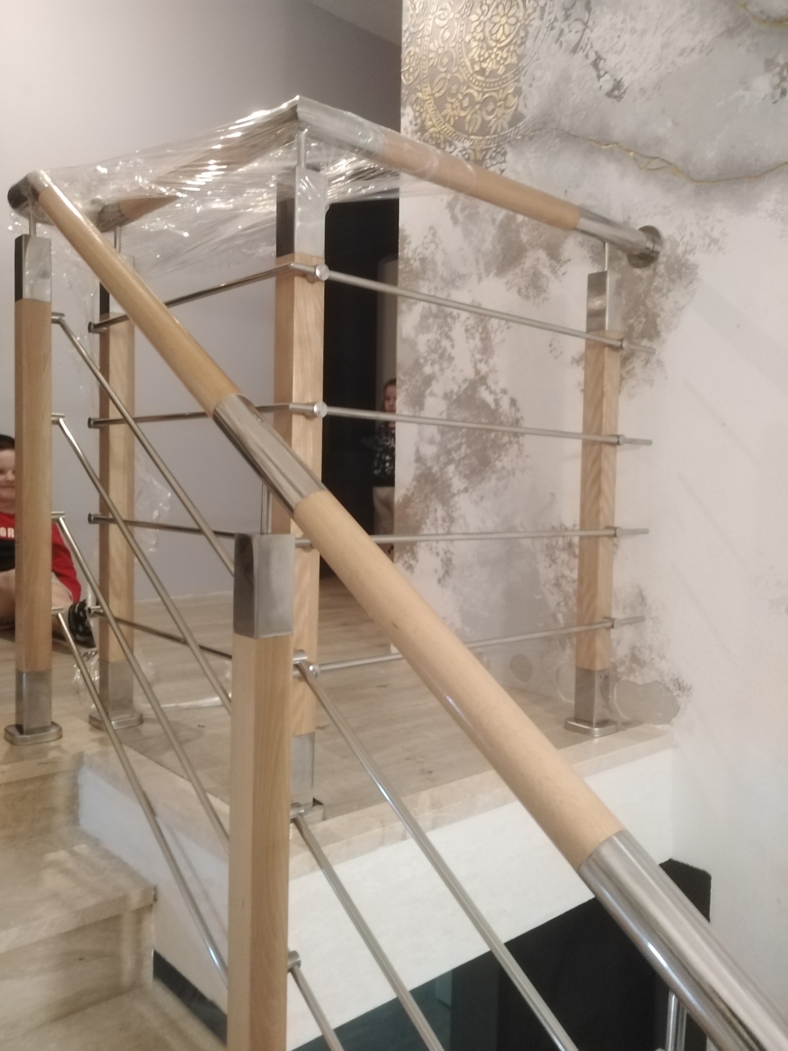 Nowoczesna balustrada schodowa z drewnianymi słupkami i stalowymi elementami wypełniającymi, częściowo owinięta folią ochronną, zamontowana na marmurowych schodach, z dekoracyjną tapetą w tle.