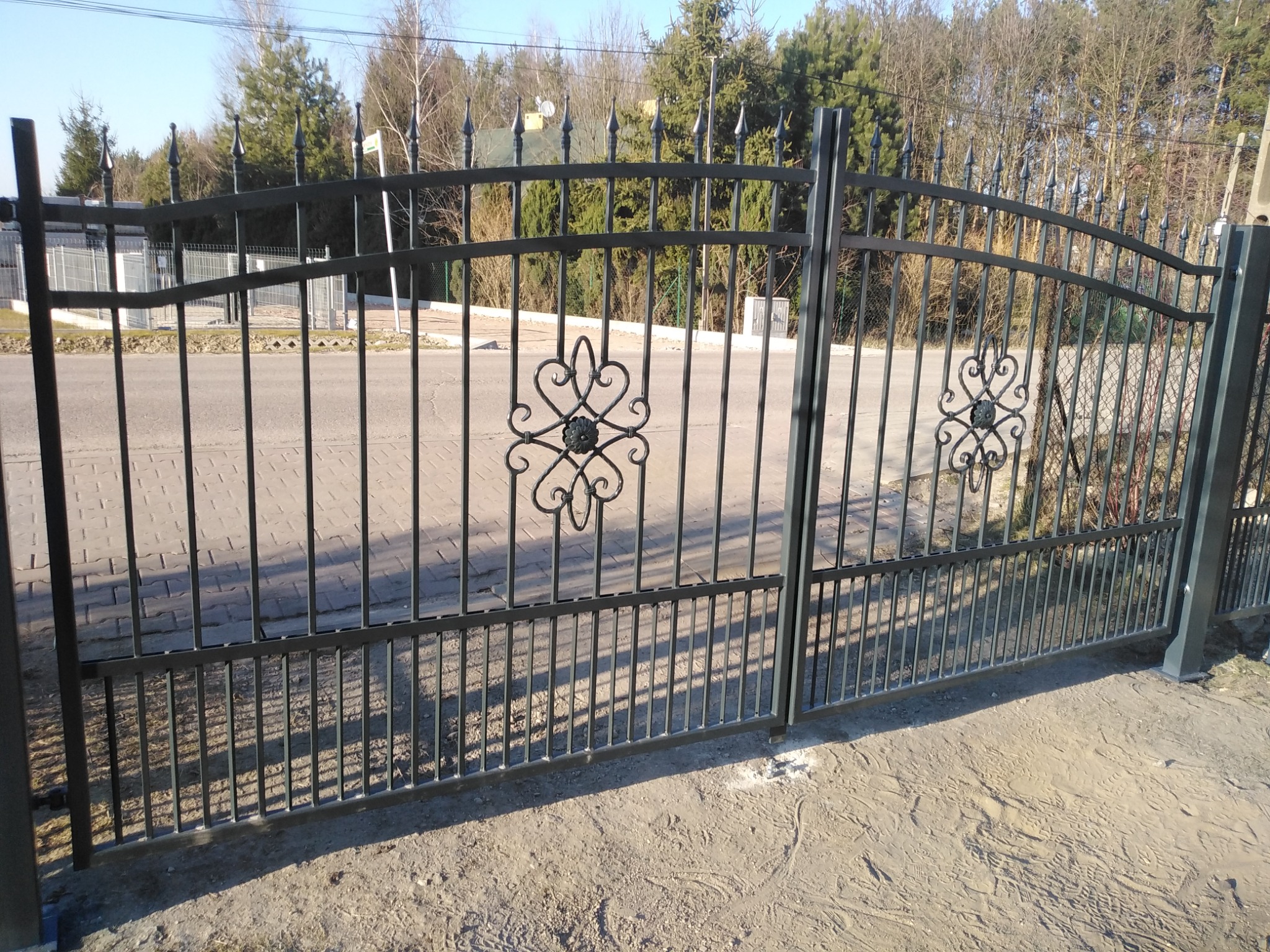 Czarna, metalowa brama wjazdowa z ozdobnymi elementami w kształcie serc i kwiatów, widoczna z bliska na tle ulicy i drzew.