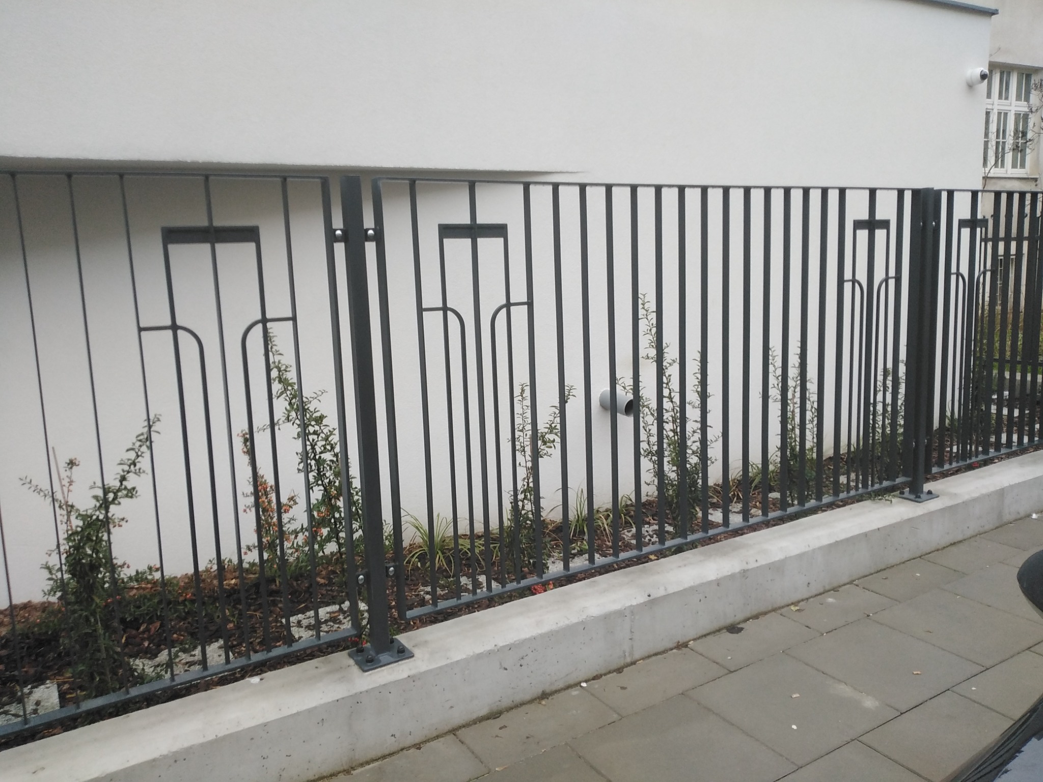 Ciemnoszare metalowe ogrodzenie z pionowych prętów z ozdobnymi elementami, zamontowane na betonowej podmurówce, z roślinnością w tle i fragmentem chodnika na pierwszym planie.