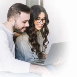 Uśmiechnięta para przegląda oferty na laptopie, kobieta w okularach i koszuli, mężczyzna w białym swetrze, jasne tło.