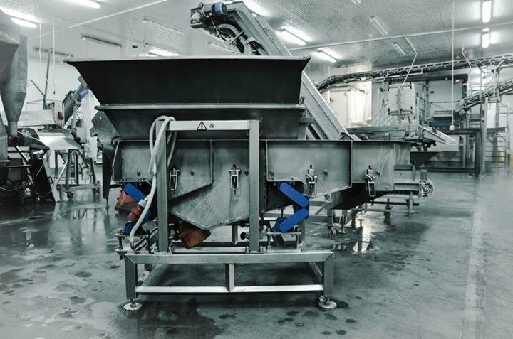 Przemysłowa linia produkcyjna z metalowymi zbiornikami i przenośnikami w sterylnych warunkach, widoczne niebieskie elementy hydrauliczne i ostrzegawcze naklejki.