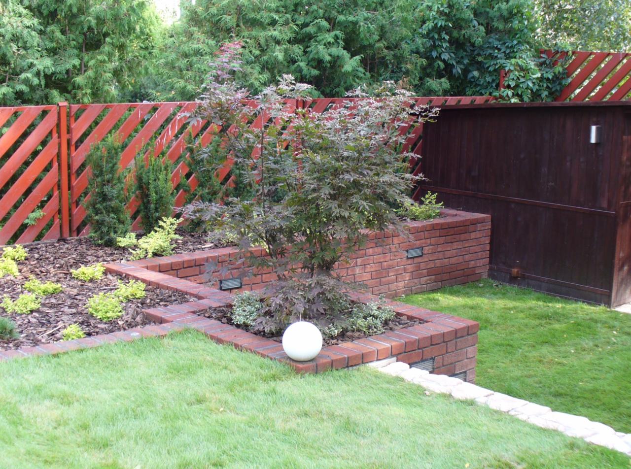Ogród z ceglanymi rabatami i ozdobnym drzewem na tle czerwonego, ukośnego ogrodzenia. W rabatach kora i niskie rośliny. Oświetlenie punktowe w murze.