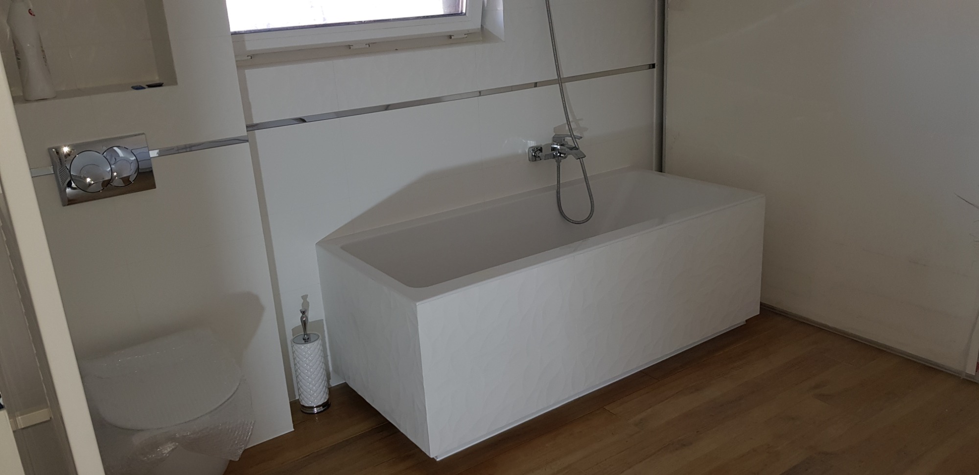 Nowoczesna łazienka z białą wanną o teksturowanej powierzchni, wiszącą toaletą z chromowanym przyciskiem spłukiwania, drewnopodobną podłogą i delikatnym oświetleniem okiennym.