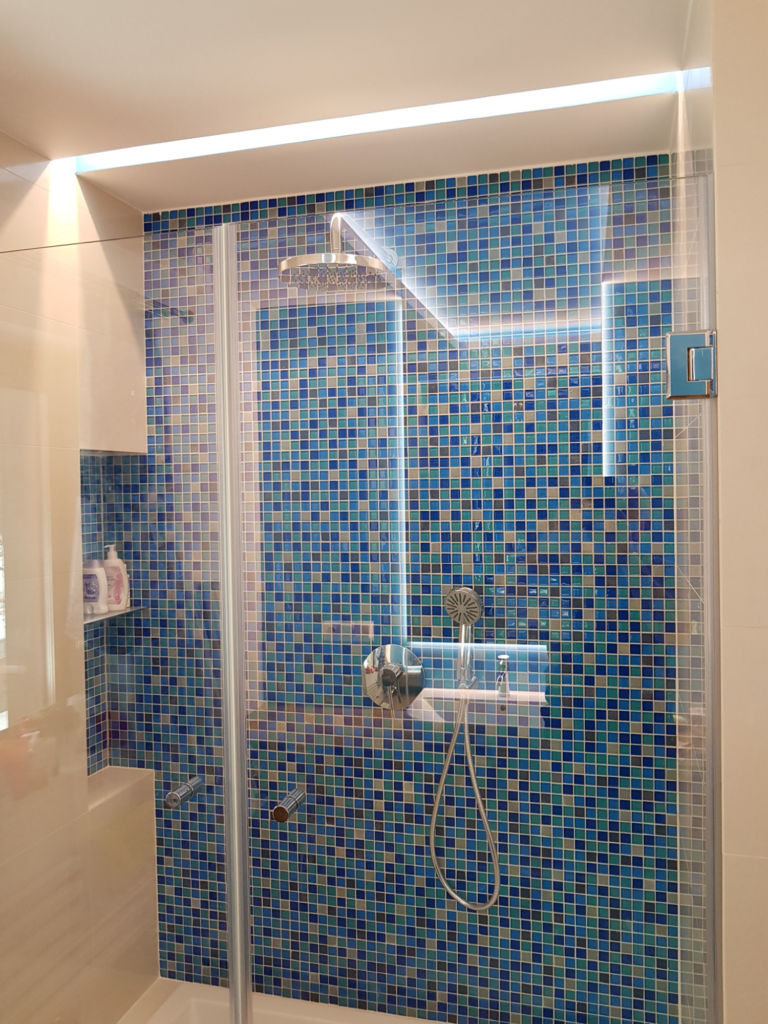 Nowoczesna kabina prysznicowa z mozaiką w odcieniach błękitu i szarości, oświetleniem LED w górnej części i dyskretną wnęką z kosmetykami.