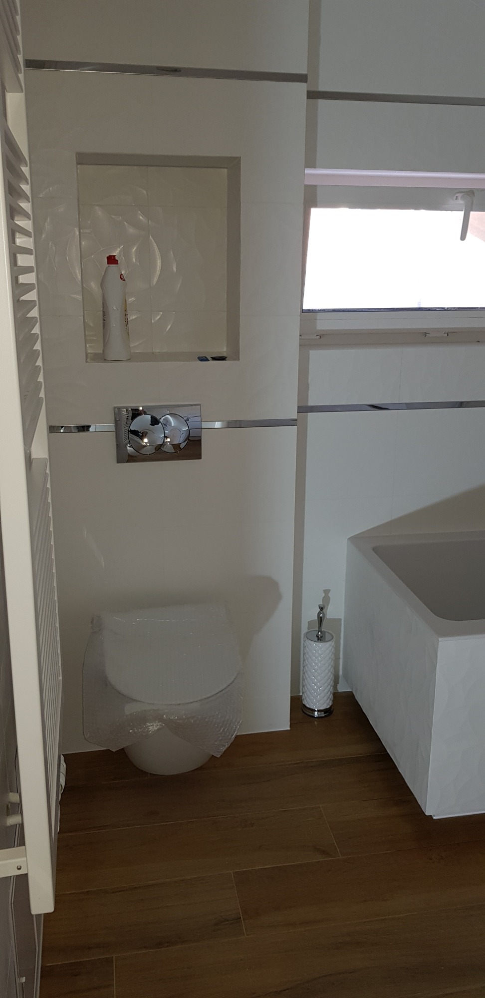 Nowoczesna łazienka z białymi płytkami, podwieszanym sedesem owiniętym folią ochronną, drewnopodobną podłogą i prostokątną wanną z panelami dekoracyjnymi.