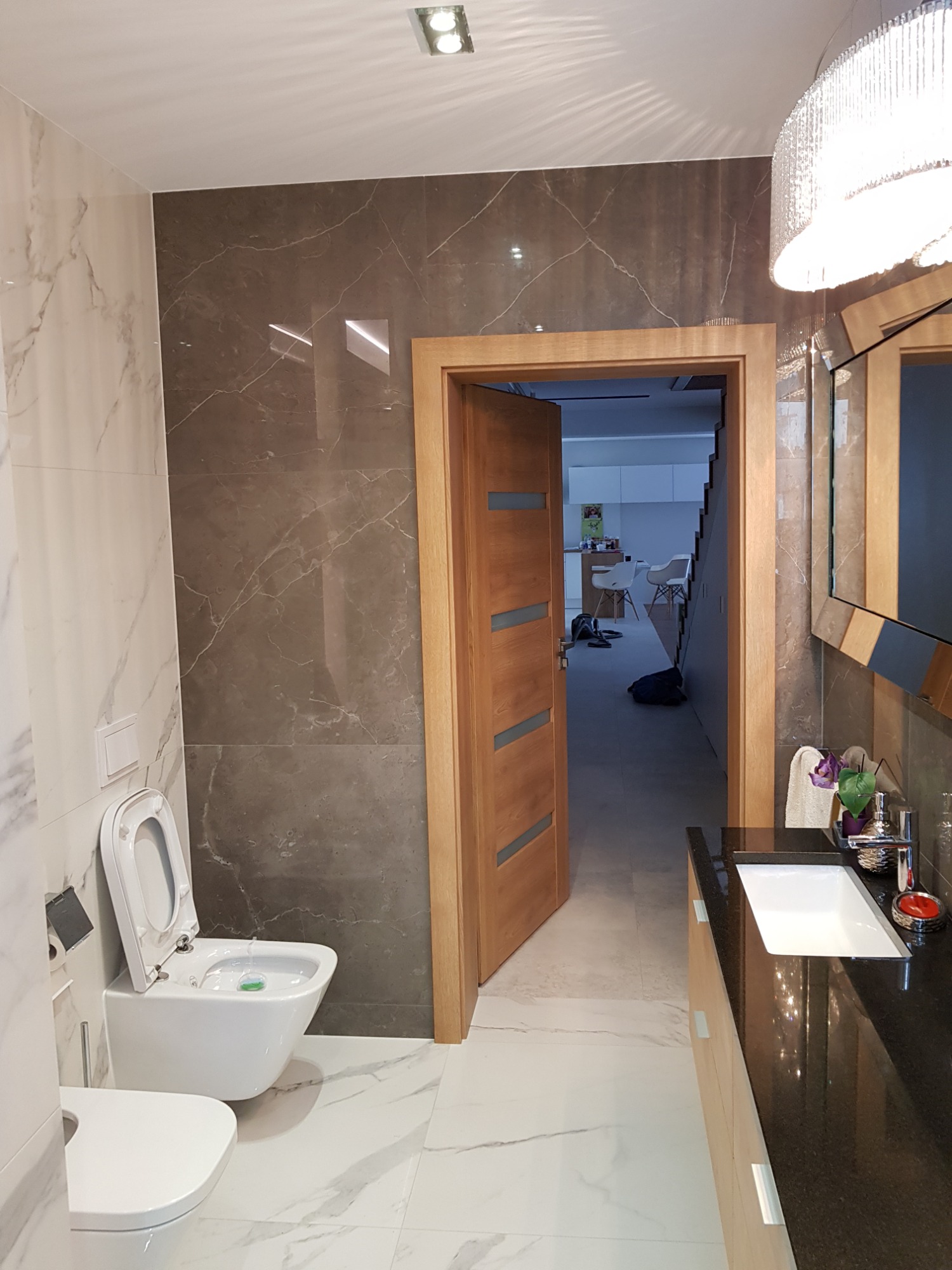 Nowoczesna łazienka z marmurowymi płytkami, podwieszaną toaletą z zielonym odświeżaczem, czarnym blatem z umywalką i kryształowym żyrandolem, widok na otwarte drzwi do dalszej części domu.