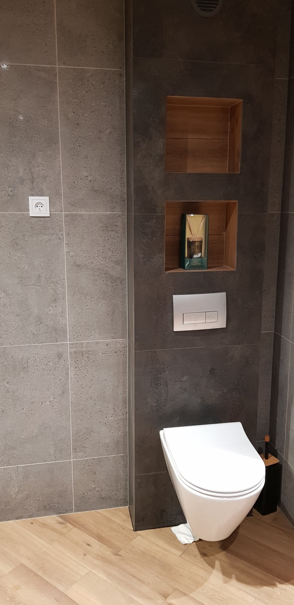 Nowoczesna toaleta podwieszana w łazience wykończonej szarymi płytkami, z drewnianymi wnękami w ścianie i drewnianą podłogą, widoczny fragment papieru toaletowego na podłodze.