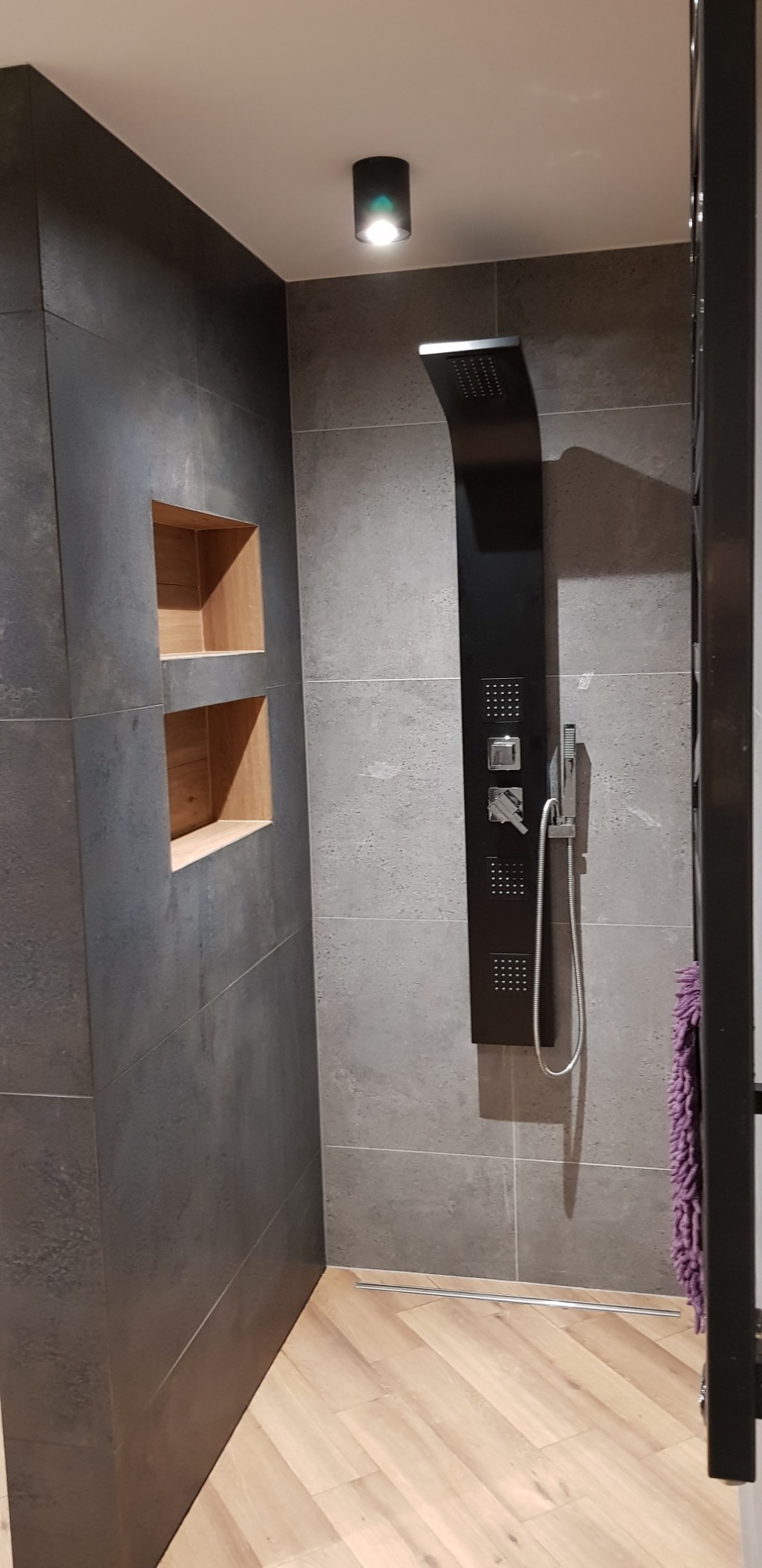 Nowoczesna łazienka z szarą płytką, drewnianą podłogą i czarnym panelem prysznicowym z deszczownicą oraz wnękami w ścianie.