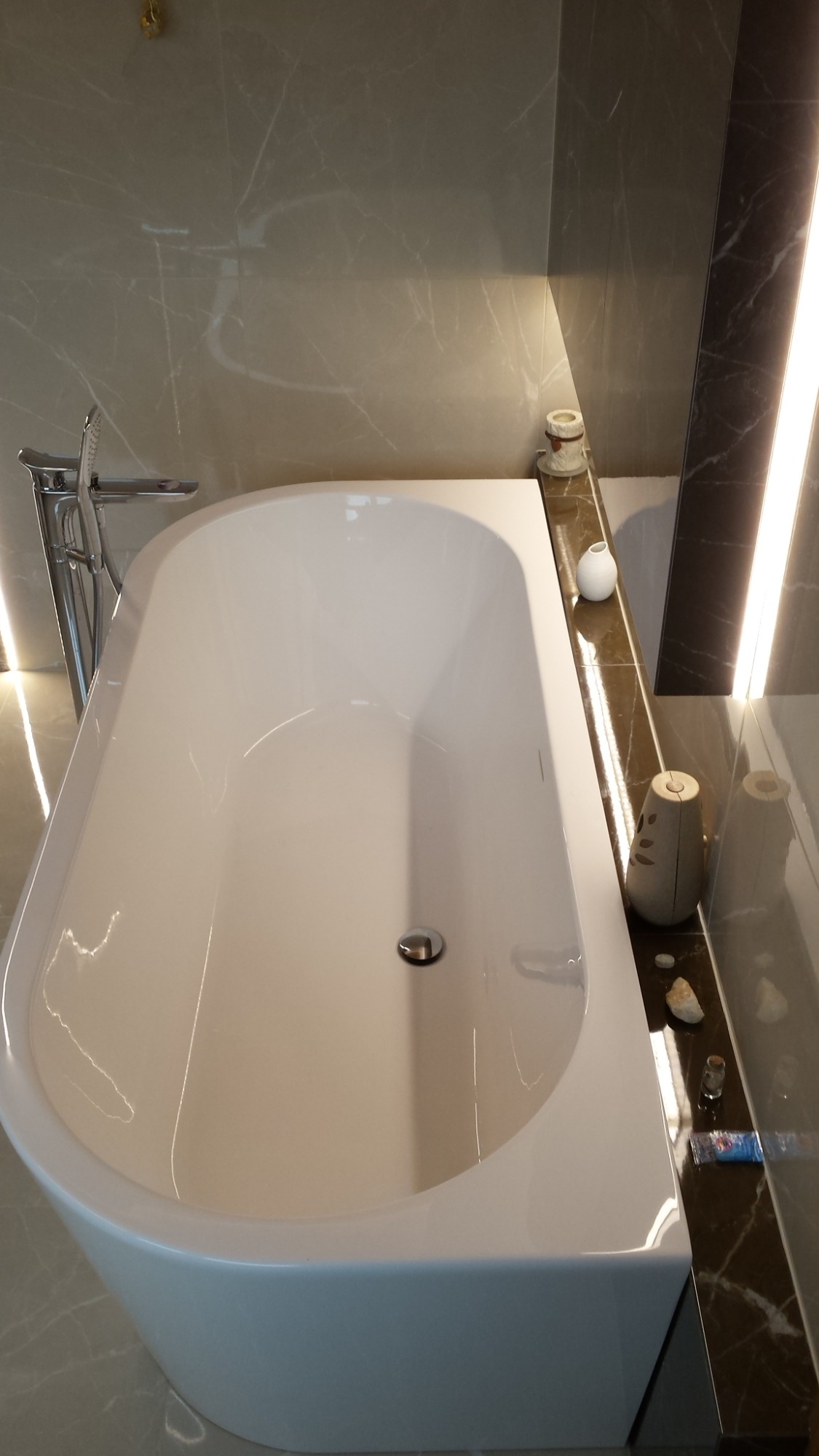 Biała, wolnostojąca wanna z chromowaną armaturą, umieszczona w łazience wykończonej płytkami imitującymi marmur, z dekoracyjnymi elementami na półce.