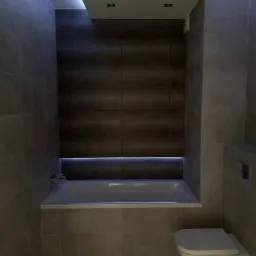 Nowoczesna łazienka z podświetlaną wanną w zabudowie i wiszącą toaletą, wykończona płytkami w odcieniach szarości i brązu, z oświetleniem LED na suficie i pod wanną.