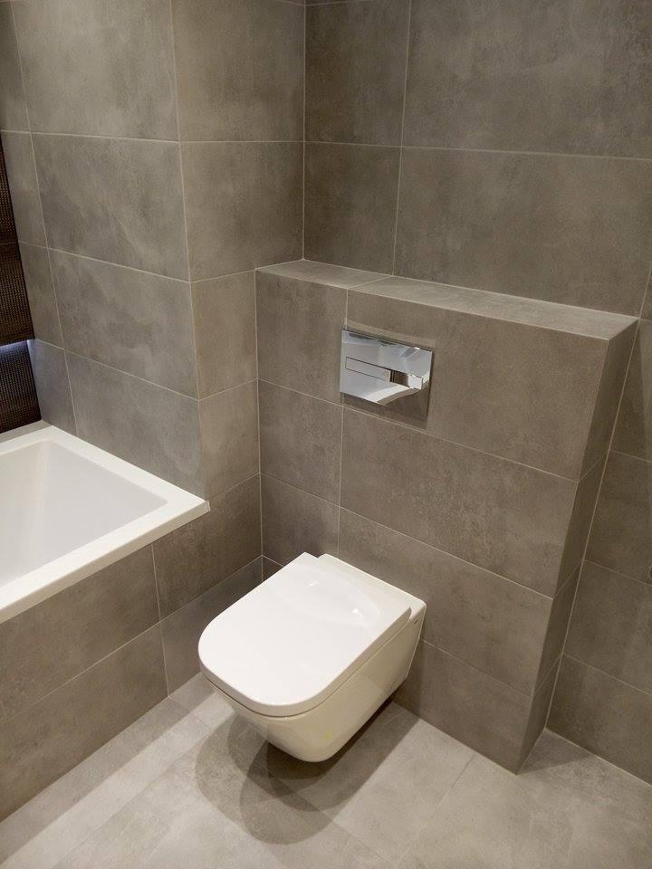 Nowoczesna łazienka z szarymi płytkami wielkoformatowymi, podwieszana toaleta z chromowanym przyciskiem spłukującym, fragment wanny po lewej stronie.