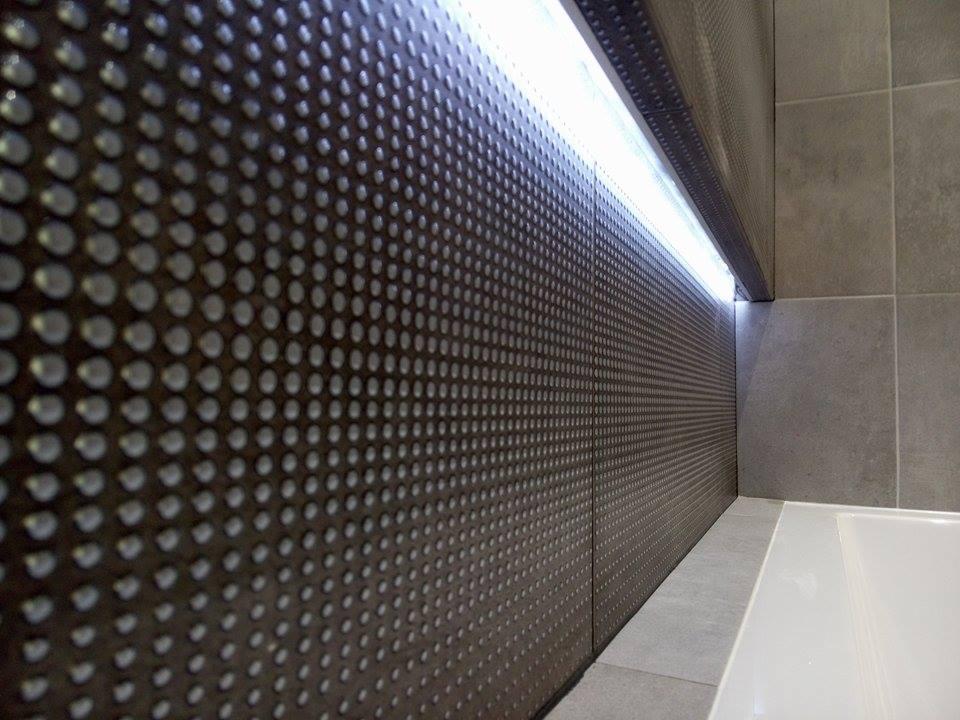 Ściana wykończona perforowanymi panelami w ciemnym kolorze, podświetlona listwą LED, sąsiadująca z szarą płytką i fragmentem białej wanny.
