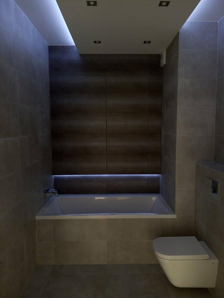 Nowoczesna łazienka z podświetlaną wanną w zabudowie i wiszącą toaletą, wykończona płytkami w odcieniach szarości i brązu, z oświetleniem LED na suficie i pod wanną.