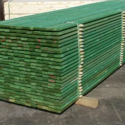Stos impregnowanych na zielono desek tarcicy ułożonych warstwami, zabezpieczonych sznurkiem, z widocznymi znakami identyfikacyjnymi na niektórych deskach.