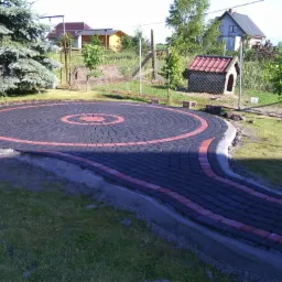 Okrągły plac z kostki brukowej w kolorze grafitowym z czerwonymi akcentami, otoczony zielonym trawnikiem i drzewami. Widoczny fragment ścieżki z kostki.