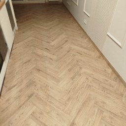 Jasna, drewniana podłoga ułożona w jodełkę francuską, świeżo po montażu. Widoczne listwy przypodłogowe i fragment ściany z dekoracyjnymi panelami.