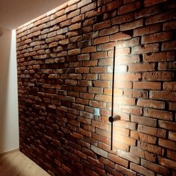 Ceglana ściana z oświetleniem LED wzdłuż krawędzi sufitu i minimalistyczną lampą ścienną. Dekoracyjne wykończenie wnętrza z efektem starej cegły.