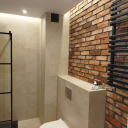 REMONTY I WYKOŃCZENIA DOMÓW I MIESZKAŃ Arkadiusz Ruszkowski - Nowoczesna łazienka z toaletą podwieszaną, beżowymi płytkami i ścianą z czerwonej cegły. Grzejnik drabinkowy w kolorze czarnym. Oświetlenie punktowe.
