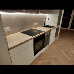 REMONTY I WYKOŃCZENIA DOMÓW I MIESZKAŃ Arkadiusz Ruszkowski - Nowoczesna kuchnia z białymi szafkami, drewnianym blatem i czarną armaturą. Uwagę przykuwa parkiet ułożony w jodełkę francuską i subtelne oświetlenie LED pod szafkami.