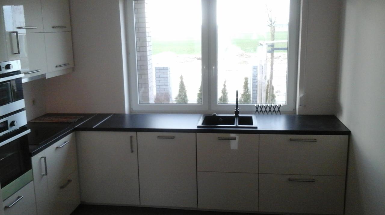 Jasna kuchnia z czarnym blatem i zlewozmywakiem, widok na okno z iglakami w tle.