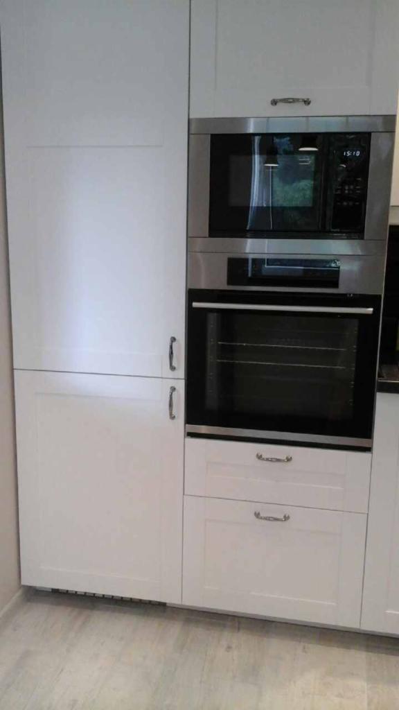 Biała zabudowa kuchenna z wbudowanym piekarnikiem i mikrofalówką ze stali nierdzewnej, widoczne dwie szuflady z metalowymi uchwytami.