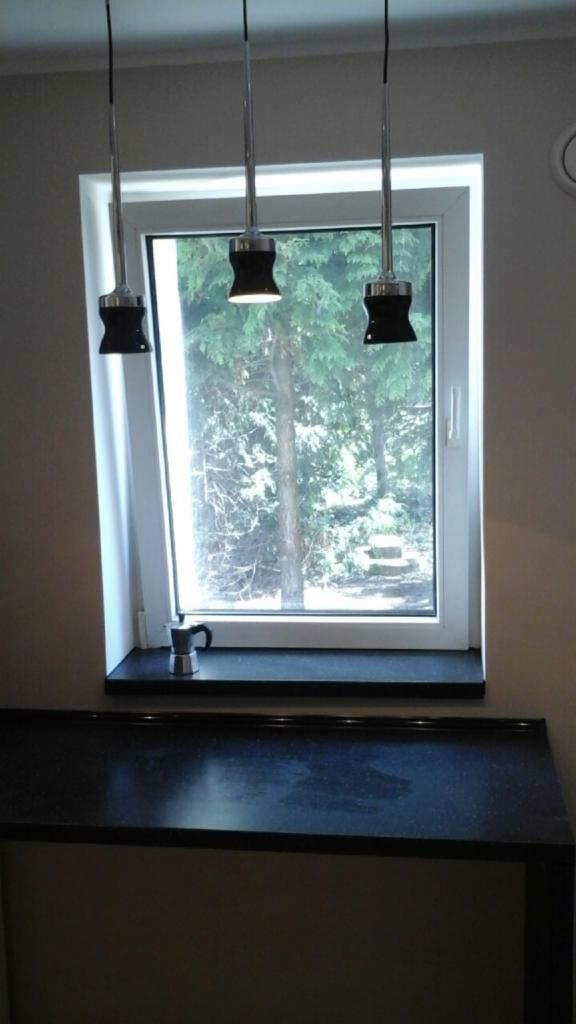 Nowoczesna kuchnia z czarnym blatem, oknem z widokiem na zieleń i trzema designerskimi lampami wiszącymi nad oknem, z małą kawiarką na parapecie.