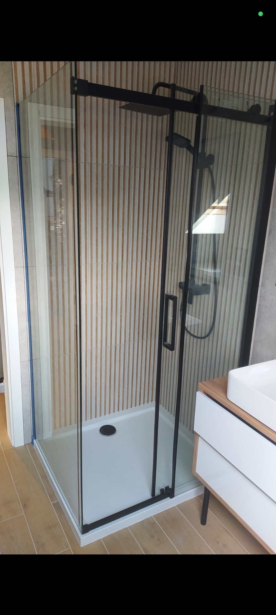 Nowoczesna kabina prysznicowa z czarnymi profilami i deszczownicą, na tle ściany z płytek imitujących drewniane lamele. Umywalka z szafką w rogu kadru.