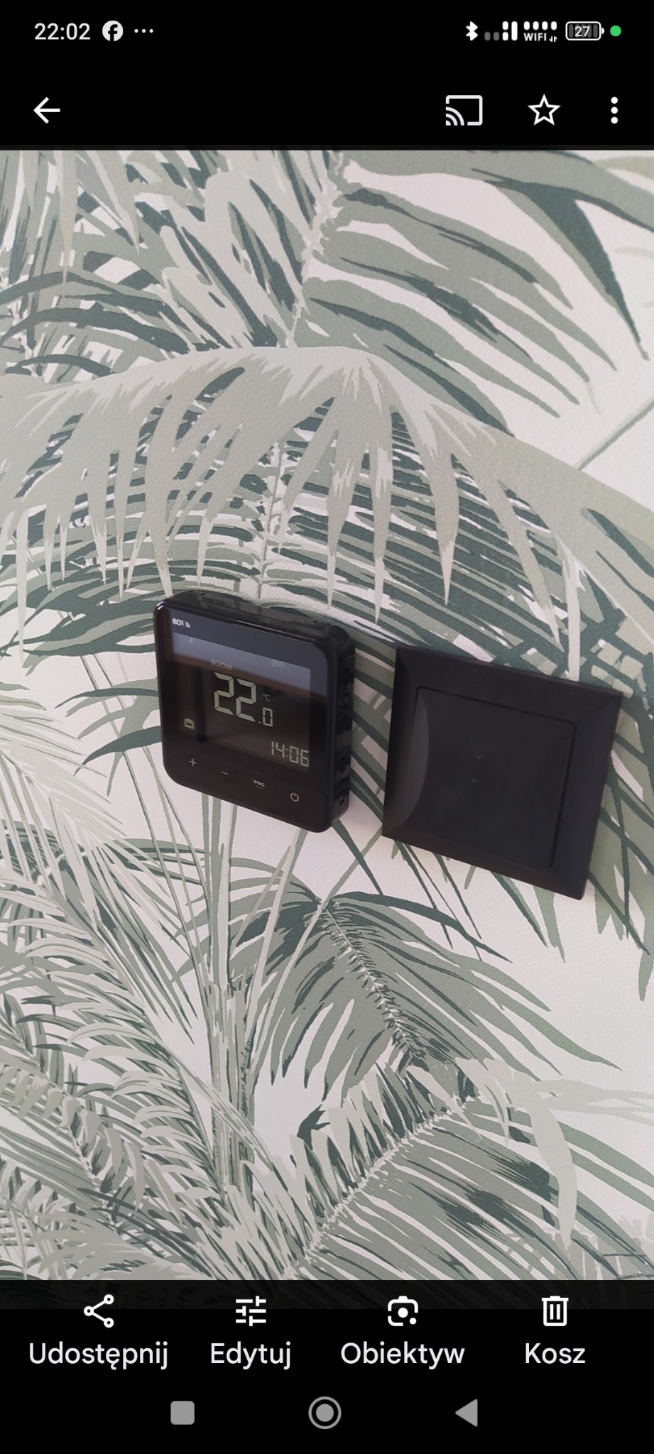 Czarny termostat i włącznik światła na ścianie z tapetą w palmy. Termostat pokazuje 22 stopnie Celsjusza i godzinę 14:06. Nowoczesny design.